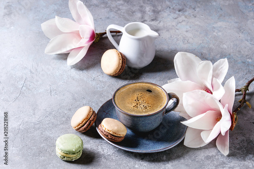 Blue cup of black espresso coffee with french dessert macaroons, cream and spring flowers magnolia branches over grey texture background. Top view, space.