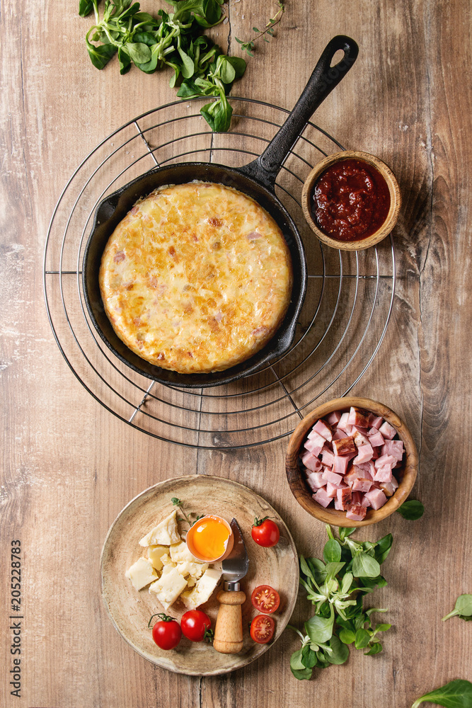 Spanish potato omelette tortilla with bacon served in cast-iron pan with sauce and ingredients above over wooden background. Top view, space. Spanish potato omelette tortilla with bacon served in cast-iron pan with sauce and ingredients above over wooden background. Top view, space.