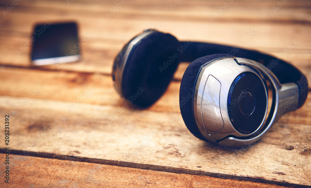 A 45 degree view of a set of wireless headphones and mobile phone lying on a rustic wooden table outside. Styling and grain effect added to image. A 45 degree view of a set of wireless headphones and mobile phone lying on a rustic wooden table outside. Styling and grain effect added to image.