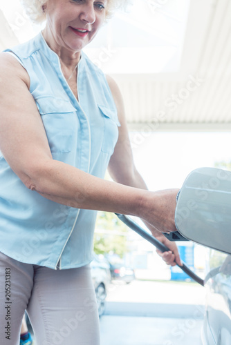 Low-angle view portrait of an active senior woman smiling while filling up the gas tank of her car at the station in summer