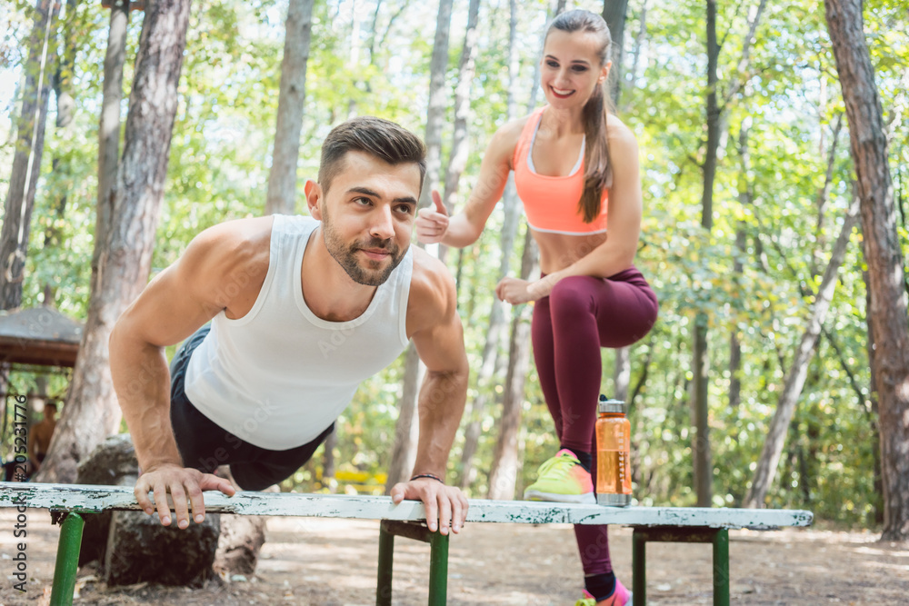 Sporty man doing push-up in an outdoor gym, his girlfriend is watching him 