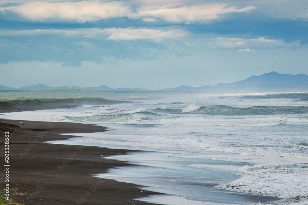 Halaktyr beach. Kamchatka. Russian federation. Dark almost black color sand beach of Pacific ocean. Stone mountains and yellow grass are on a background. Light blue sky