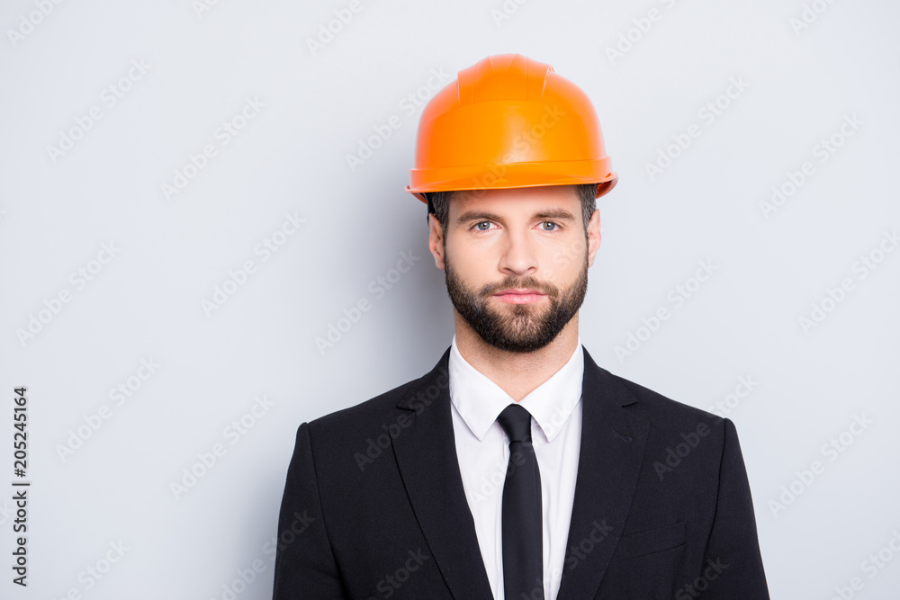 Portrait of concentrated, thoughtful architector with stubble in orange safety helmet, hardhat and black tux with tie, looking at camera, isolated on grey background Portrait of concentrated, thoughtful architector with stubble in orange safety helmet, hardhat and black tux with tie, looking at camera, isolated on grey background