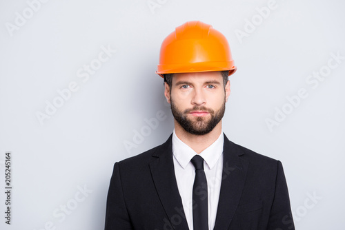 Portrait of concentrated, thoughtful architector with stubble in orange safety helmet, hardhat and black tux with tie, looking at camera, isolated on grey background © deagreez