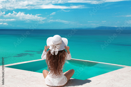 Carefree Woman relaxing in infinity swimming pool looking at view. Luxury resort. Beautiful destination summer vactions. Back view of traveller girl in beach hat and white bikini relax on Maldives. photo