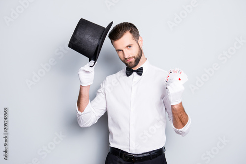 Portrait of virile harsh illusionist with stubble and modern hairstyle, holding tophat and set of cards in hands, looking at camera, isolated in grey background
