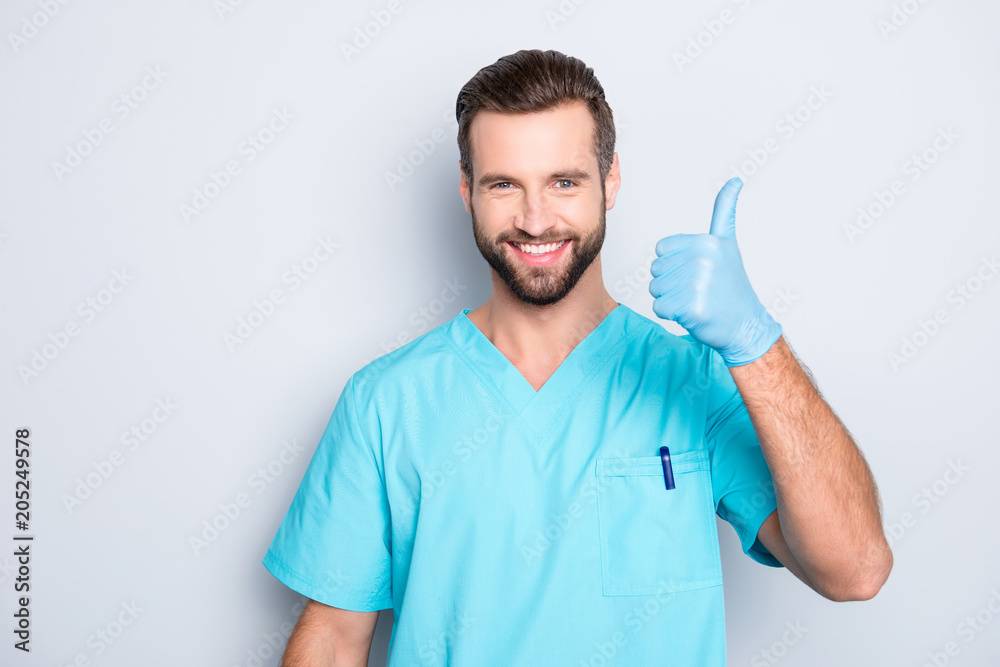 Portrait of joyful positive medic with stubble in lab blue uniform looking at camera showing thumb up sign with hand, finger, isolated on grey background, advertisement concept Portrait of joyful positive medic with stubble in lab blue uniform looking at camera showing thumb up sign with hand, finger, isolated on grey background, advertisement concept