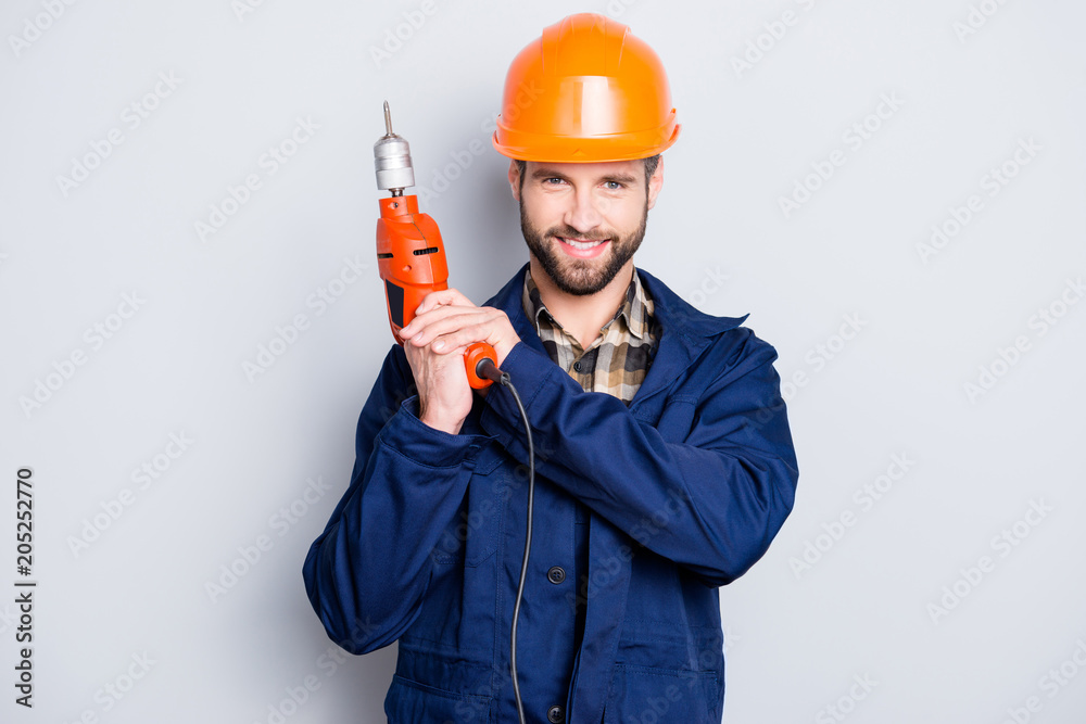 Portrait of joyful positive repairer in protective hard hat with stubble showing, having equipment in arm isolated on grey background Portrait of joyful positive repairer in protective hard hat with stubble showing, having equipment in arm isolated on grey background