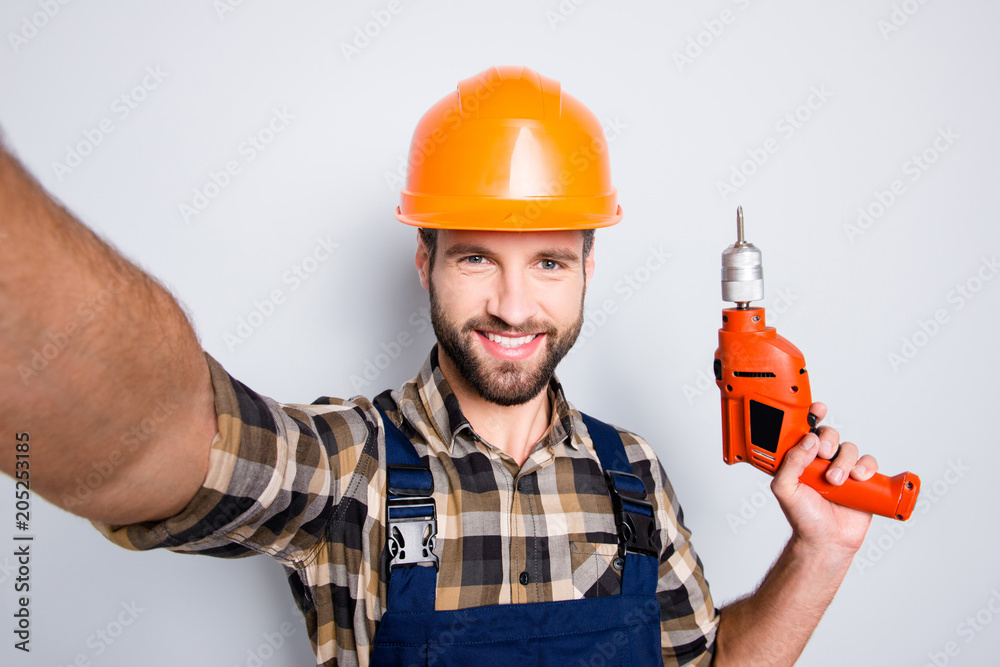 Self portrait of cheerful joyful repairer in protective hard hat shooting selfie on front camera having equipment in hand isolated on grey background. Leisure fun concept Self portrait of cheerful joyful repairer in protective hard hat shooting selfie on front camera having equipment in hand isolated on grey background. Leisure fun concept