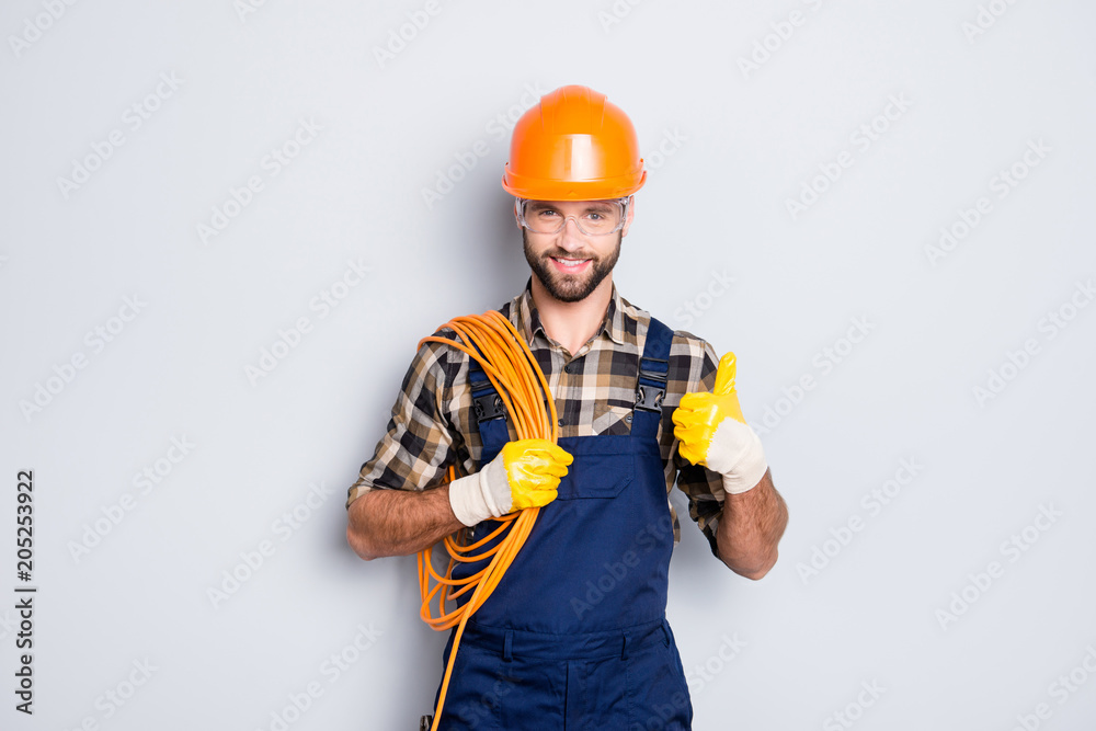 Portrait of handsome joyful electrician in hardhat, overall, shirt with bristle, holding rolled wires on shoulder, showing thumb up recommend approve sign over grey background Portrait of handsome joyful electrician in hardhat, overall, shirt with bristle, holding rolled wires on shoulder, showing thumb up recommend approve sign over grey background