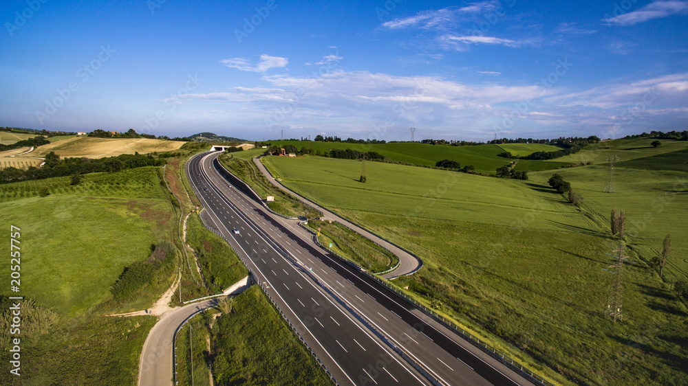 Italia, Maggio 2018 - Vista aerea dell'autostrada con automobili e camion che attraversa le colline con i campi di grano non ancora maturo