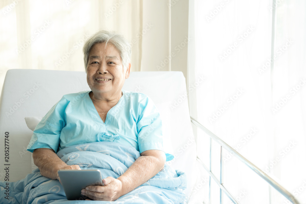 Elderly patient using tablet in bed. Elderly chinese woman in hospital bed using tablet to connect with her relatives. Connected world concept. Elderly patient using tablet in bed. Elderly chinese woman in hospital bed using tablet to connect with her relatives. Connected world concept.