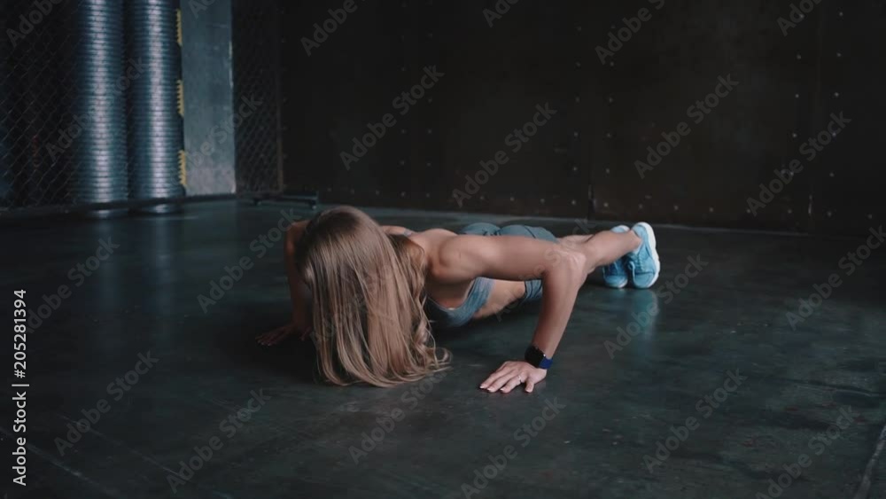 Slider shot of an attractive young woman wearing a gray sports bra, shorts and smartwatch doing pushups in a gym. Slow motion medium shot