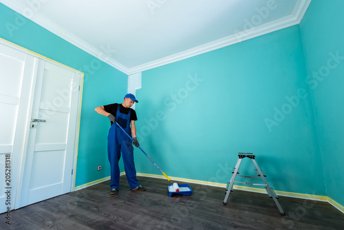 Man in a working overall is painting the wall in white color