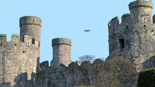UFO sighting, Flying saucer hovering over Conwy castle, metallic reflective aircraft rotating and moving, realistic reconstruction of real UFO sighting, blue sky background.