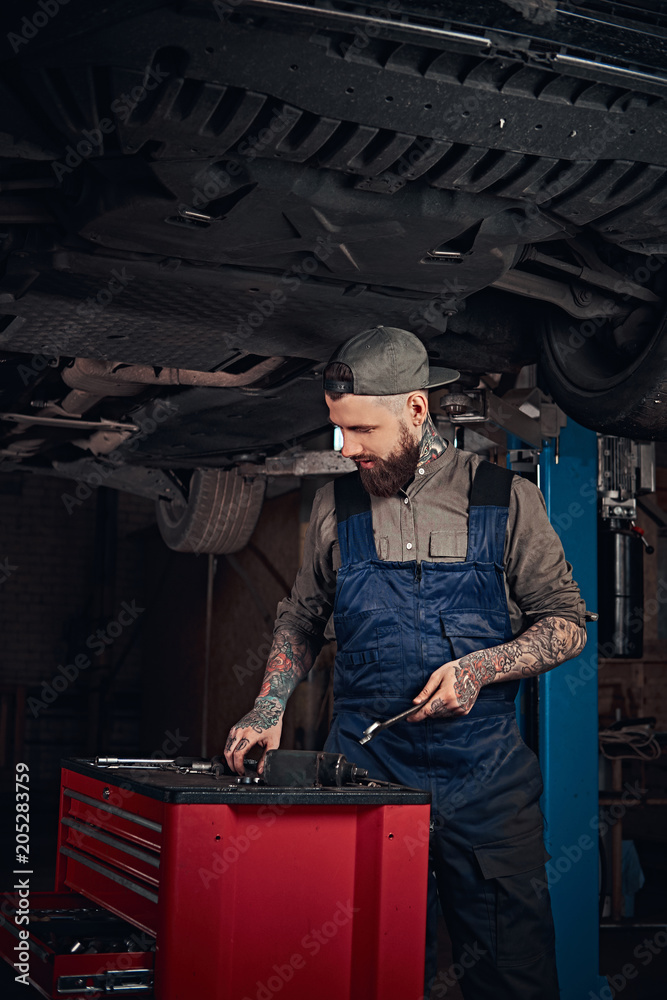 Auto mechanic in a uniform, working on a workbench while standing under lifting car in a repair garage. Auto mechanic in a uniform, working on a workbench while standing under lifting car in a repair garage.