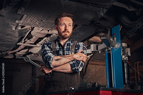 Mechanic crossed hands while standing under lifting car in a repair garage.