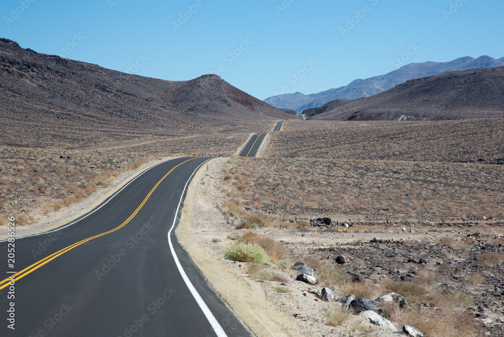 Eine unendliche lange und geschlängelte aphaltierte Strasse mit gelben Mittelstreifen zieht sich durch die trockene amerikanische Wüste bei blauem Himmel