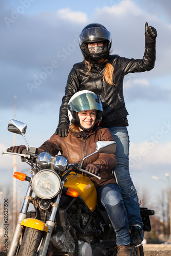 Two joyful European women driving motorcycle, girl standing behind with thumb up