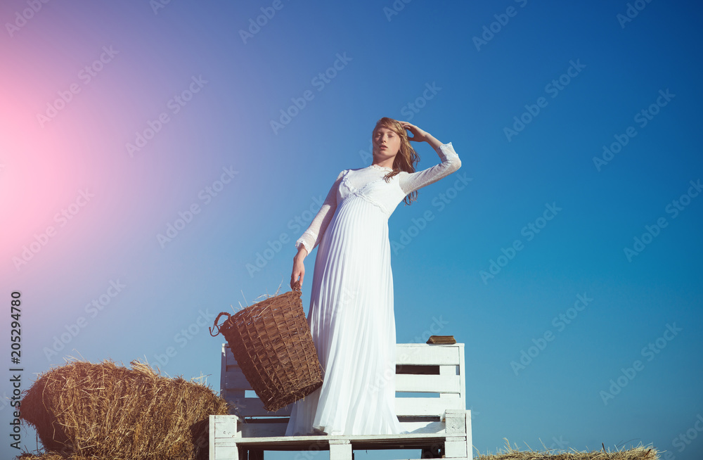 Fashion model on blue sky. Albino girl hold wicker basket with hay on sunny  outdoor picnic. Woman bride in wedding dress on wooden bench. Sexy woman  with long blond hair Stock Photo