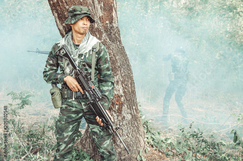 Soldier standing behind a tree ready to attack. Chinese male soldier standing behind a large tree looking around for his ememy and covering his friend. Smoke effect.
