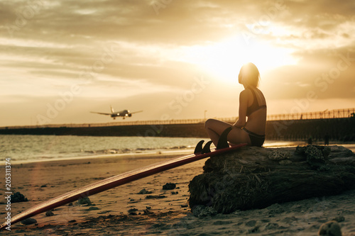woman sitting on rock with surfboard on beach at sunset with airplane in sky