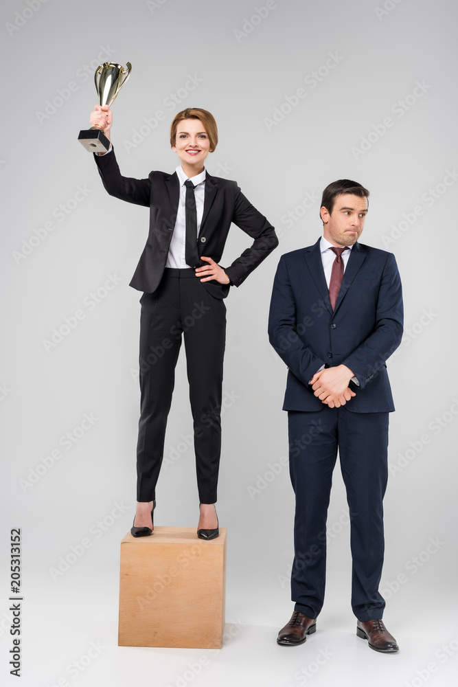 happy businesswoman with trophy cup standing on podium, upset colleague standing near, isolated on grey happy businesswoman with trophy cup standing on podium, upset colleague standing near, isolated on grey