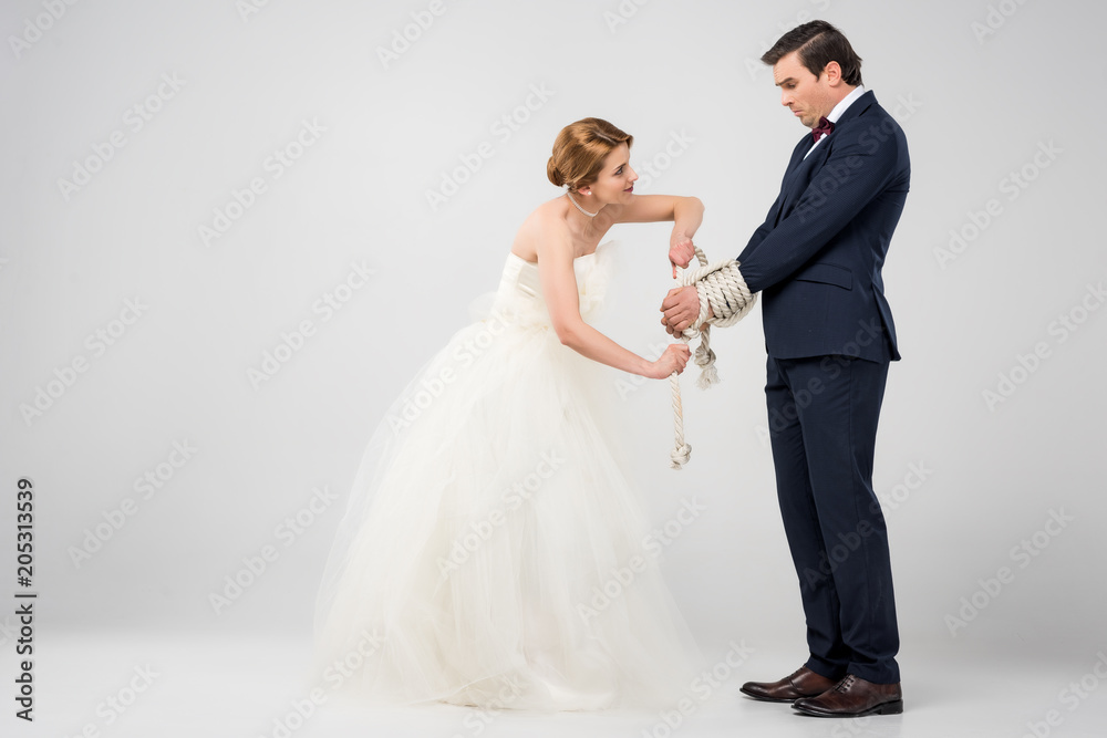 bride in wedding dress bounding groom with rope, isolated on grey, feminism concept
