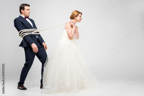 bride in wedding dress pulling groom bound with rope, isolated on grey, feminism concept