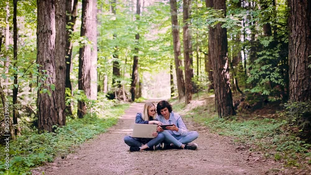 Young Couple Using Laptop And Tablet While Sitting In Forest Young Couple Using Laptop And Tablet While Sitting In Forest