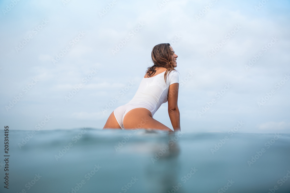 attractive young woman in wet white swimsuit sitting on surfboard in ocean