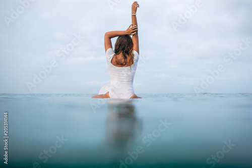 rear view of young woman in wet white swimsuit sitting on surfboard in ocean