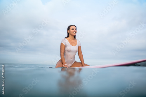 smiling attractive woman in white wet swimsuit sitting on surfboard in ocean during summer vacation