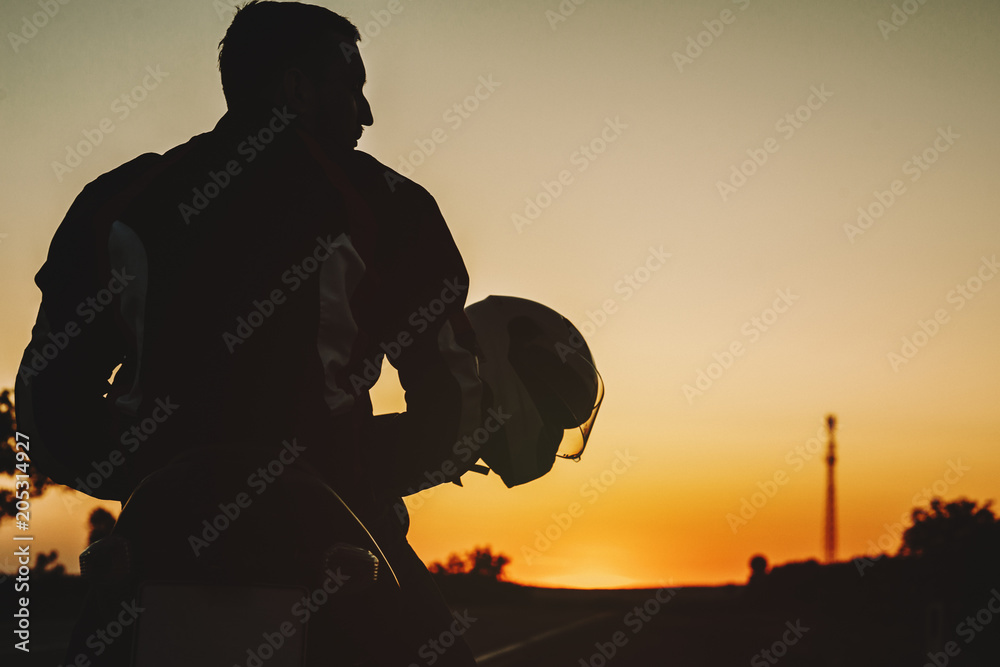 Back view of a caucasian young biker sitting on his motorcycle and looking away against sunset light.
