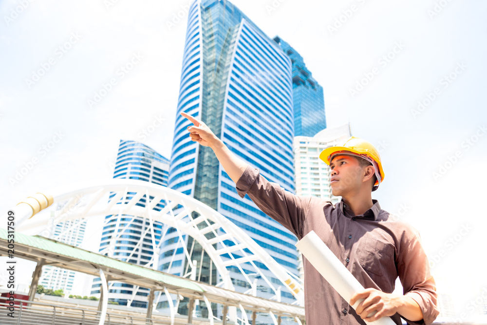 Handsome worker or engineer man is pointing finger, looking to copy space. Employee hold blueprint. Asian guy look determined and confident. He wear hard hat for safety at workplace. city background Handsome worker or engineer man is pointing finger, looking to copy space. Employee hold blueprint. Asian guy look determined and confident. He wear hard hat for safety at workplace. city background