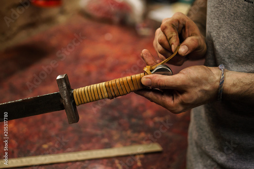 Smith makes leather winding of sword. Man is working in workshop. He fixes leather band on hilt of sword.