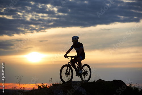 Robust and strong man wearing sportswear and helmet sitting on his bike and posing on hill. Cyclist enjoying great view of sunset. Concept of motivation, recreation and healthy activities. © anatoliy_gleb