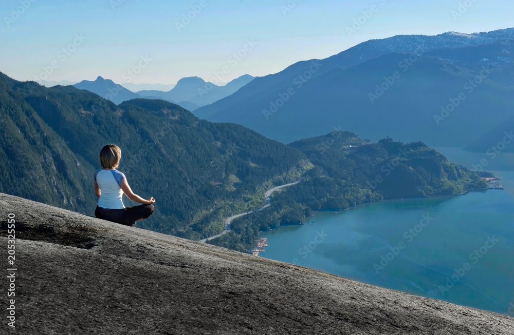 Outdoor yoga retreat. Woman in lotus pose meditating on mountain top with beautiful views. Whistler. Stawamus Chief Peak. British Columbia. Canada.