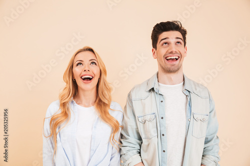 Portrait of attractive young people man and woman in basic clothing looking upwards with happy smile, isolated over beige background