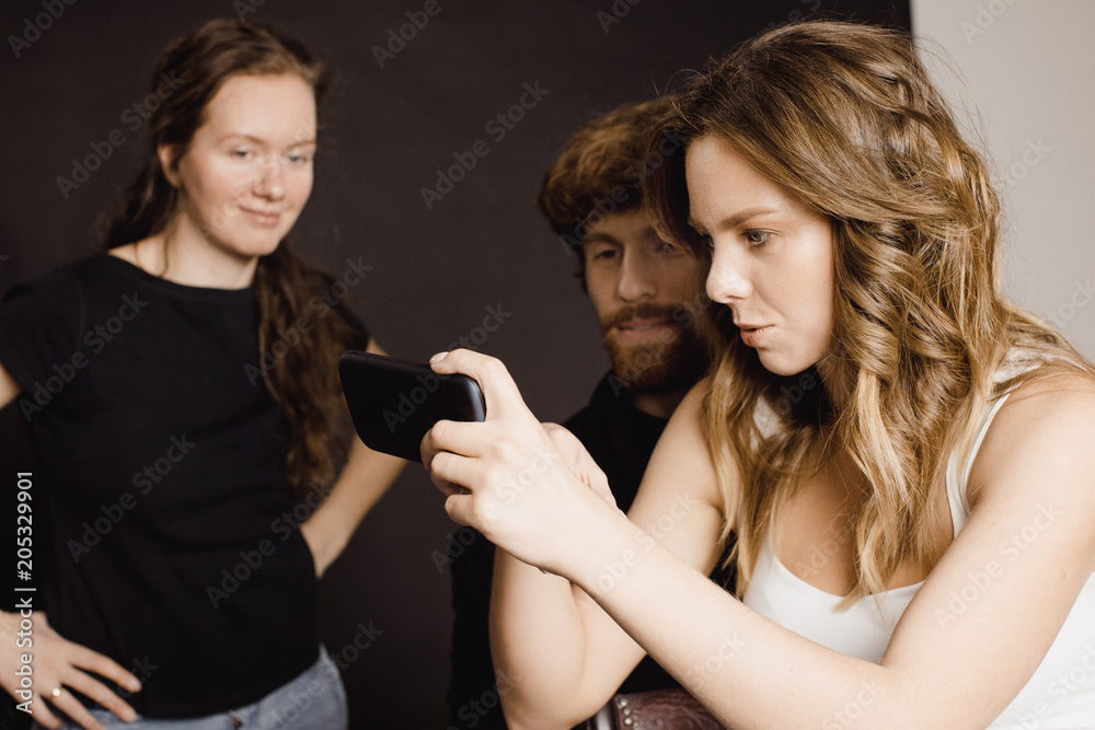 Three people in casual outfits holding smartphone and looking at photos on it while standing in studio. Three people in casual outfits holding smartphone and looking at photos on it while standing in studio.