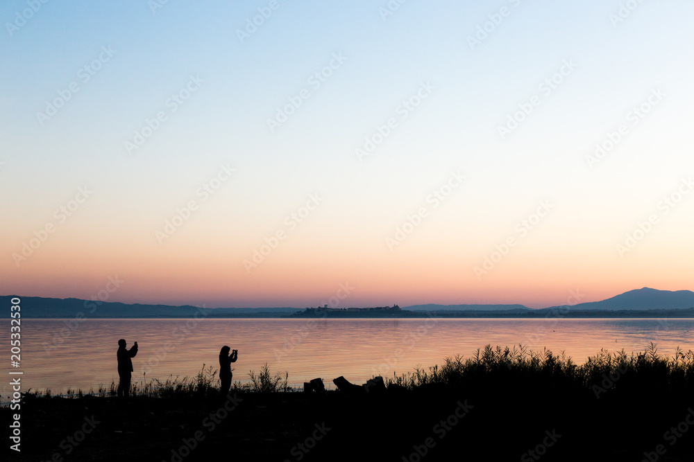 Silhouettes of a couple on a lake shore taking photos of sunset 