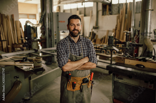Portrait of a smiling young woodworker standing with his arms crossed by a bench saw in his workshop full of carpentry equipment