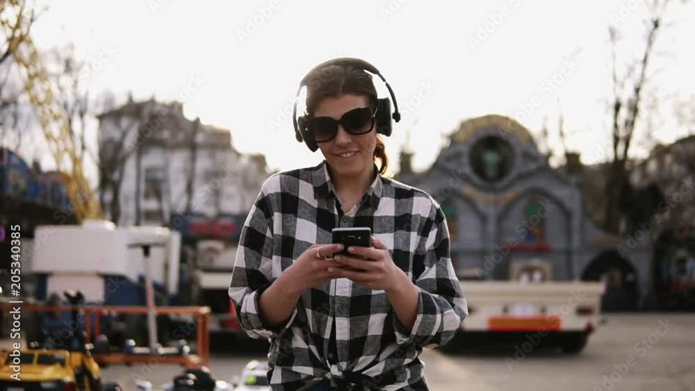 Trendy, brunette in sunglasses walking on the street and listen to the music in headphones. Dances. Holding mobile in hands and searching for something. Sunny day Trendy, brunette in sunglasses walking on the street and listen to the music in headphones. Dances. Holding mobile in hands and searching for something. Sunny day