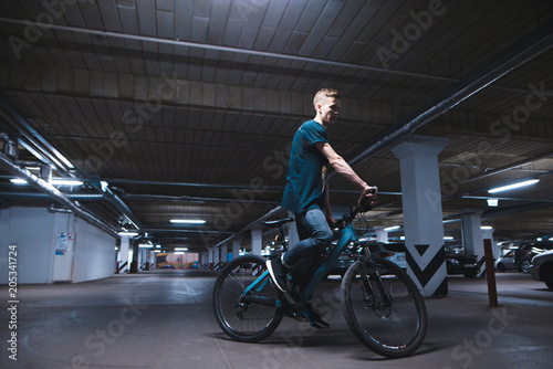 Fototapeta Naklejka Na Ścianę i Meble -  Portrait of a cyclist in an underground parking lot for cars. A man rides a bicycle by parking