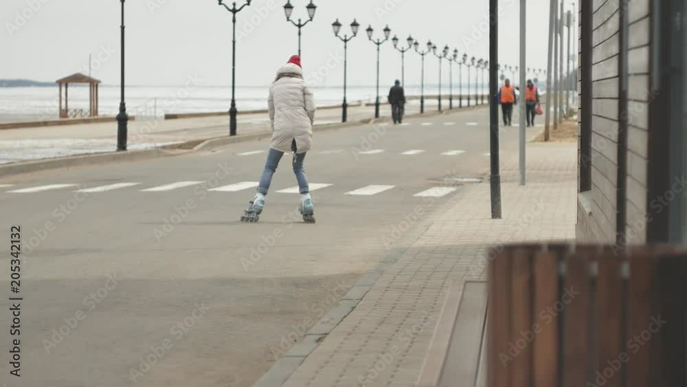 Young beautiful woman in a red hat, wearing sporty warm clothes and rollers, riding on the road on the coast Young beautiful woman in a red hat, wearing sporty warm clothes and rollers, riding on the road on the coast