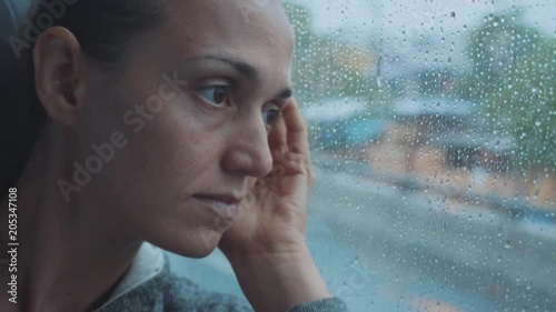 Portrait of young sad woman looking out the wet window, while travelling by bus.