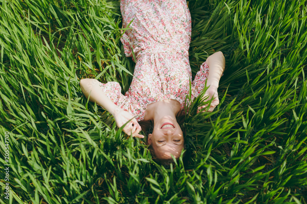 Young smiling pretty woman in light patterned dress lying on grass spreading hands looking up resting in sunny weather in field on bright green background. Spring nature. Lifestyle, leisure concept. Young smiling pretty woman in light patterned dress lying on grass spreading hands looking up resting in sunny weather in field on bright green background. Spring nature. Lifestyle, leisure concept.