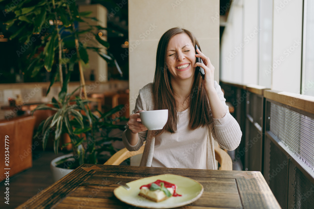 Young woman sitting alone in coffee shop at table with cup of cappuccino, cake, relaxing in restaurant during free time. Young female talking on mobile phone, having rest in cafe. Lifestyle concept.