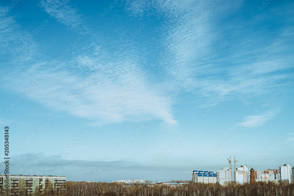 Spring cityscape with high-rise building, under construction, cranes under blue cloudy sky on foreground from grove. Trails of airplane in sky.