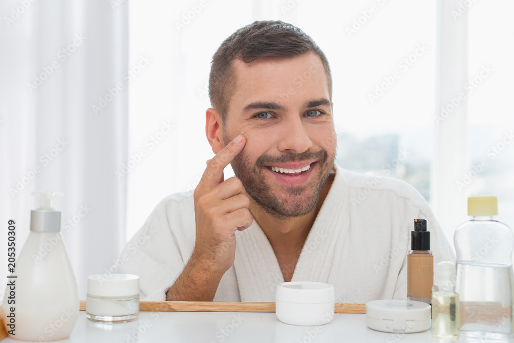 Positive mood. Delighted nice man touching his face while applying facial cream